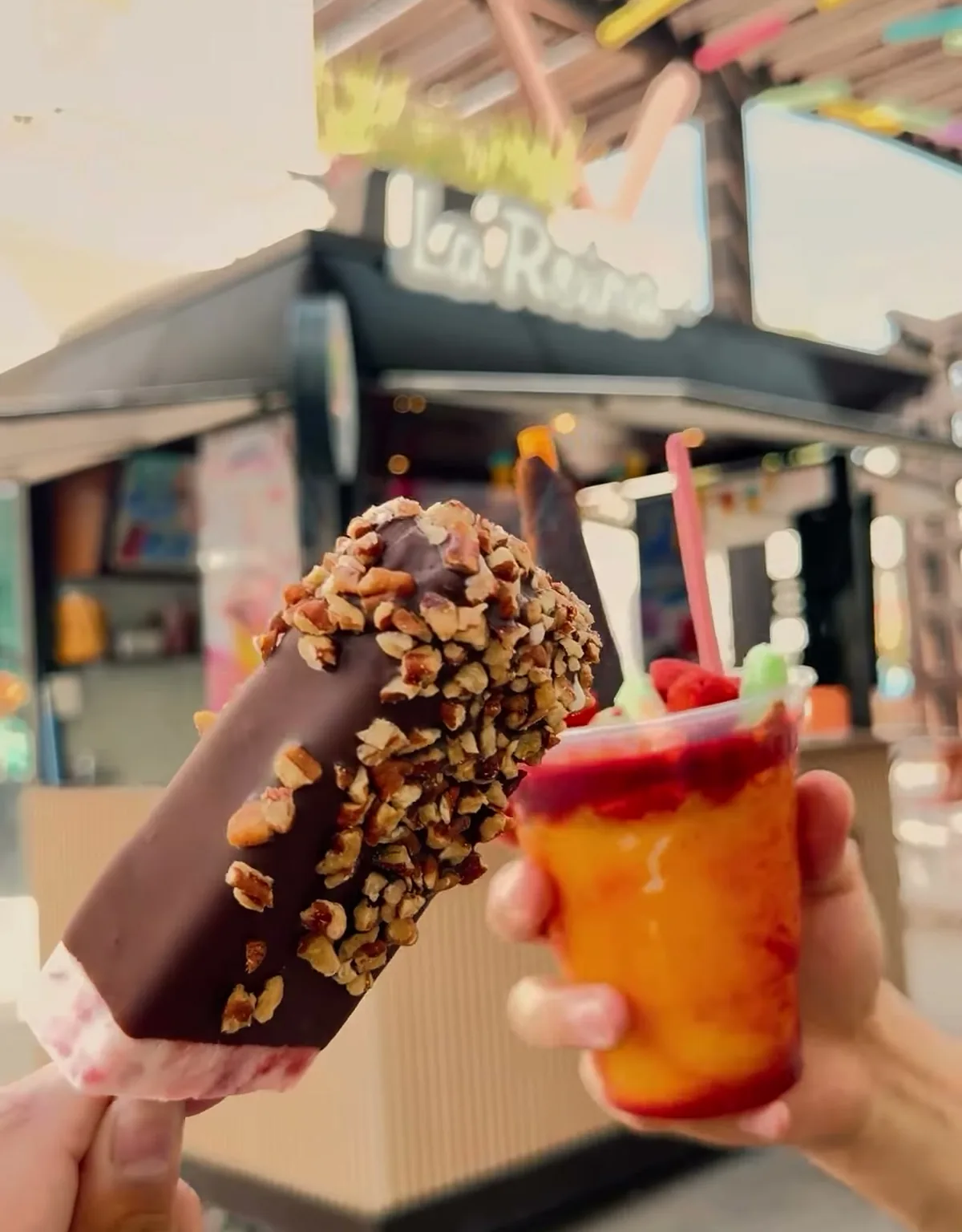 Una paleta cubierta de chocolate y nuez y una mangonada de mango, sostenidas frente al local de La Reina.